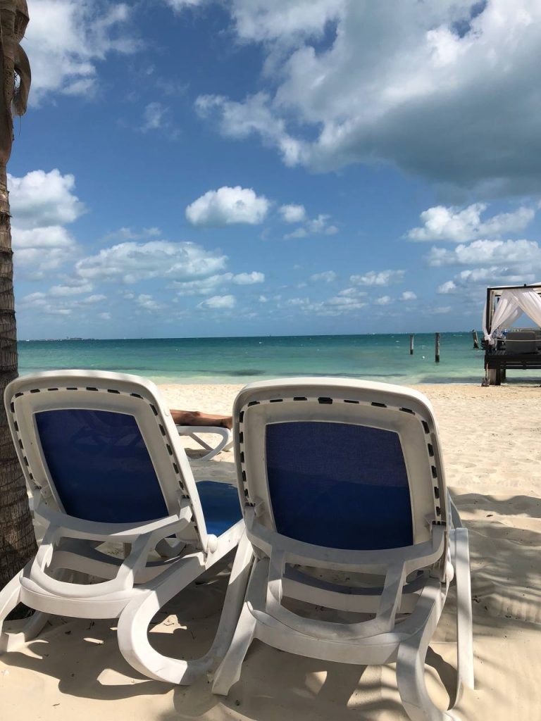 Two white lounge chairs on a sandy beach overlooking a calm ocean under a bright blue sky